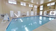 This indoor aquatic facility is centered around a generously sized swimming pool with crystal-clear blue water, making it a standout feature of the space. The pool is bordered by a clean, light-colored tile surround and is equipped with a metal entry ladder for easy access, offering a safe and functional environment for swimmers of all levels.

The room's architecture is defined by its elevated ceiling height, which creates an open and airy atmosphere. A row of clerestory windows lines the upper portion of the walls on multiple sides, allowing natural light to pour in and reflect off the water's surface, enhancing the bright and refreshing ambiance throughout the day.

Along one wall, a series of individual changing rooms with white doors are evenly spaced, providing private accommodations for users. The lower portion of the walls features a tile wainscoting in a neutral stone tone, adding a clean and durable finish that complements the overall design. A simple wooden bench is positioned poolside for convenience.

The warm, neutral-toned walls and smooth ceiling contribute to a calm and inviting environment. The single-floor layout ensures easy accessibility throughout the facility. Overall, this space is thoughtfully designed to serve as a functional and comfortable indoor swimming destination.