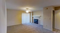 This single-floor residential unit offers a practical and comfortable layout with a warm, neutral aesthetic throughout. The living area serves as the centerpiece of the space, featuring wall-to-wall carpeting that extends through the unit, providing a soft and cohesive feel underfoot. A standout feature of the living area is the tile-surround fireplace, which adds both visual interest and a cozy focal point to the room. A ceiling fan with a light fixture overhead provides ventilation and ambient lighting, while a set of vertical blinds covers a full sliding glass door, allowing for natural light and potential outdoor access. The walls are finished in a warm beige tone that complements the neutral carpeting throughout. A ceiling-mounted HVAC vent ensures climate control across the unit. A hallway connects the living space to additional rooms, offering a sense of privacy and separation between living and other functional areas of the unit. The overall ambiance is clean, understated, and move-in ready, with a simple design that provides a versatile canvas for personalization.
