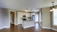 This single-floor space showcases an open-concept layout centered around a well-appointed kitchen. The kitchen features crisp white cabinetry that extends to the upper walls, providing generous storage. A central island with a dark countertop serves as both a workspace and a focal point of the room, illuminated by two pendant lights that hang at varying heights, adding a layered lighting effect. Stainless steel appliances, including a built-in microwave, gas range, and French door refrigerator, complement the modern aesthetic. The countertops carry a consistent dark tone that contrasts cleanly with the white cabinetry below.

The flooring throughout is a warm hardwood-style material that adds visual continuity across the open space. Neutral greige-toned walls keep the interior feeling cohesive and understated. A door with a glass panel at the far end of the kitchen area allows natural light to filter into the space, brightening the interior. The open layout connects the kitchen to adjacent living areas, making the overall space feel welcoming and functional. Recessed and pendant lighting ensures the space is well-lit throughout.