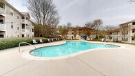 Located at 1200 Lanier Mill Circle in Oakwood, Georgia, this outdoor amenity area serves as a central gathering space within a well-maintained residential community. The centerpiece of the space is a generously sized freeform swimming pool with clear, blue water, offering both shallow and deeper sections clearly marked for safety. The pool is surrounded by a broad concrete sundeck equipped with lounge chairs, providing ample space for relaxation and recreation.

A dedicated poolside amenity building is positioned at the far end of the pool deck, featuring a teal-accented entrance and serving as a functional support facility for the area. The pool area is fully enclosed by black wrought-iron fencing, ensuring security while maintaining an open, airy feel. Manicured hedges and mature trees line the perimeter, adding natural greenery and privacy to the space.

The surrounding multi-story residential buildings, clad in neutral siding with blue shutters and featuring private balconies, frame the pool area and contribute to the cohesive community aesthetic. A well-kept landscaped entry path with rounded shrubs and a gated access point leads into the pool enclosure. The overall ambiance is welcoming and well-organized, making this outdoor space a functional and attractive communal amenity for residents of the Lanier Mill Circle community in Oakwood, GA.