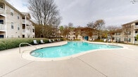 Located at 1200 Lanier Mill Circle in Oakwood, Georgia, this outdoor amenity area serves as a central gathering space within a well-maintained residential community. The centerpiece of the space is a generously sized freeform swimming pool with clear, blue water, offering both shallow and deeper sections clearly marked for safety. The pool is surrounded by a broad concrete sundeck equipped with lounge chairs, providing ample space for relaxation and recreation.

A dedicated poolside amenity building is positioned at the far end of the pool deck, featuring a teal-accented entrance and serving as a functional support facility for the area. The pool area is fully enclosed by black wrought-iron fencing, ensuring security while maintaining an open, airy feel. Manicured hedges and mature trees line the perimeter, adding natural greenery and privacy to the space.

The surrounding multi-story residential buildings, clad in neutral siding with blue shutters and featuring private balconies, frame the pool area and contribute to the cohesive community aesthetic. A well-kept landscaped entry path with rounded shrubs and a gated access point leads into the pool enclosure. The overall ambiance is welcoming and well-organized, making this outdoor space a functional and attractive communal amenity for residents of the Lanier Mill Circle community in Oakwood, GA.