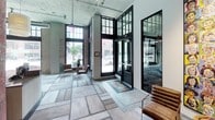 This single-floor space presents a thoughtfully designed commercial lobby that seamlessly merges industrial character with contemporary aesthetics. The entrance is defined by soaring ceilings, floor-to-ceiling black-framed windows, and large-format stone-look tile flooring that floods the space with natural light and street views. Exposed concrete ceilings and original brick walls reinforce the building's industrial heritage throughout.

At the heart of the lobby sits a curved reception desk topped with a smooth gray surface, accompanied by a striking terracotta-red structural column that adds architectural interest. Colorful wall art and curated artwork pieces are displayed throughout, contributing to a vibrant, gallery-like atmosphere. Leather sling chairs provide casual seating near the entrance.

A dedicated beverage station features dark wood cabinetry, a herringbone terracotta tile backsplash, a wall-mounted television, and a compact red refrigerator, creating a welcoming refresh area. Adjacent to this is a dramatically designed lounge alcove with a dark latticed ceiling, deep red padded wall panels, booth-style seating, and moody recessed lighting, offering an intimate gathering space. The overall ambiance balances energy and sophistication.