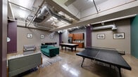 This industrial-chic amenity lounge offers a dynamic, open-concept layout designed for recreation and relaxation. The space is defined by its raw architectural character, including exposed ductwork and mechanicals overhead, polished concrete floors, and bold accent columns painted in deep eggplant purple — all of which lend an urban loft aesthetic throughout.

The main game area is generously proportioned, housing both a full-size billiard table and a ping-pong table, providing ample room for play without feeling cramped. A vibrant teal feature wall serves as a striking backdrop in the game zone, complemented by framed artwork that adds a curated, gallery-like touch. A bookshelf stocked with reading materials sits nearby, offering a quieter diversion.

The lounge area is thoughtfully furnished with a tufted emerald-green sofa, mid-century modern accent chairs, and a patterned area rug that anchors the seating zone. A wall-mounted flat-screen TV, dark wood media cabinet, and an articulating floor lamp create an inviting nook for unwinding. A small dining table with sleek chairs provides an additional gathering spot.

A kitchenette along the back wall features warm wood cabinetry, a stainless steel refrigerator, and a countertop workspace, ensuring convenience for social gatherings. The overall ambiance balances industrial edge with comfortable, well-appointed furnishings for a versatile communal space.