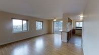 This single-floor space offers an open, airy layout with a seamless flow between the living area and kitchen. The living area is bathed in natural light from multiple windows that frame lush outdoor views, creating a welcoming and bright atmosphere throughout the day. Smooth, light-toned hardwood floors run throughout, adding warmth and a sense of continuity to the space. A ceiling fan centered in the living area provides both functional airflow and a classic finishing touch. Sliding glass doors lead directly to a private balcony, extending the living space outdoors and offering pleasant elevated views of the surrounding greenery. The kitchen is partially separated by a peninsula with dark wood cabinetry, providing ample storage and a natural visual divide between the cooking and living zones. Upper cabinetry with glass-front panels adds character to the kitchen area, while a window above the kitchen allows additional natural light. The neutral, beige-toned walls throughout provide a clean and versatile backdrop, making the space highly adaptable for a variety of furnishing styles. Overall, the space combines functional design with an inviting, light-filled ambiance well-suited for comfortable everyday living.
