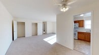 This single-floor residence at 2 Underhill Rd in Middletown, New York offers a clean, open layout filled with natural light. The main living area features neutral-toned wall-to-wall carpeting, crisp white walls, and a ceiling fan with integrated lighting, contributing to a bright and comfortable atmosphere. Two generously sized closets with white panel doors provide abundant storage directly within the living space, a practical and convenient design choice. A baseboard heating unit runs along the far wall, ensuring efficient climate control throughout the space. The open sightlines connect the living area to adjacent rooms, creating a seamless flow through the unit. A doorway visible from the main room leads further into the residence, suggesting additional living or sleeping space beyond. The adjoining kitchen is partially visible and features white appliances, including a range and microwave, set against warm wood-toned cabinetry. The overall design is straightforward and functional, with neutral finishes that offer a versatile backdrop. The combination of natural sunlight streaming through windows, ample storage, and an open floor plan makes this a practical and welcoming single-floor living space.