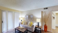 This single-floor space centers around a comfortable living area designed with a clean, neutral palette that creates a warm and inviting atmosphere. The living room features a plush, light-toned sofa accented with yellow and cream throw pillows, adding a cheerful pop of color against the soft beige walls. Two framed artworks are symmetrically displayed above the sofa, contributing to the room's balanced aesthetic. A dark-finished wood coffee table anchors the seating area over a textured area rug, while matching table lamps on either side of the sofa provide warm ambient lighting. The room is finished with a textured ceiling and includes a floor-to-ceiling vertical blind window treatment, suggesting access to generous natural light and potentially an outdoor area beyond. Hardwood-style flooring runs throughout the space, adding warmth and continuity. A decorative arrangement of tall dried branches in an amber glass vase near the entryway adds an organic, artistic touch. The overall ambiance is cozy yet polished, making the living area a functional and aesthetically pleasing centerpiece of the home.