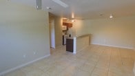 This single-floor space is designed with an open-concept layout that connects the living area and kitchen, creating a unified and functional environment. The flooring throughout is consistent light-toned ceramic tile, lending a clean, cohesive look to the space.

The kitchen is equipped with standard white appliances, including a refrigerator, electric range, and microwave. Warm-toned wood cabinetry provides ample storage, and a track lighting fixture overhead ensures the cooking area is well lit. A two-toned breakfast bar peninsula with a dark lower base and light countertop serves as a natural divider between the kitchen and the living area, offering additional seating and counter space.

The living area is spacious and open, featuring neutral beige walls and white baseboards that contribute to a bright, airy atmosphere. A ceiling fan is visible, adding to the practicality of the space. A bifold-door closet is accessible just off the main area, providing convenient storage. The overall ambiance is clean, functional, and welcoming, with a simple, straightforward design suited to everyday living.