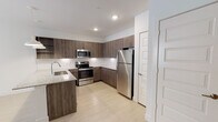 This single-floor residence showcases a contemporary design with a cohesive color palette of whites, light wood tones, and warm cabinetry finishes. The kitchen serves as a centerpiece of the space, outfitted with full-height wood-grain cabinetry providing generous storage, a granite-topped peninsula island with an undermount sink, and a full suite of stainless steel appliances including a refrigerator, range, and built-in microwave. Recessed lighting and elegant pendant lights above the island illuminate the space, complemented by a clean white tile backsplash. Light wood-look flooring runs throughout the main living areas, contributing to a bright and airy atmosphere.

The entryway is clean and functional, with light flooring and direct access to adjacent rooms. A dishwasher is conveniently integrated near the kitchen entry. The bedroom features plush neutral carpeting, a ceiling fan with integrated light, and a sliding-door closet with built-in shelving, offering practical storage. The bathroom is finished with light wood-look flooring, a full-sized soaking tub and shower combination with white tile surround, and brushed nickel fixtures. Overall, the space presents a clean, modern aesthetic well-suited for comfortable everyday living.