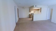 This single-floor space offers an open-concept layout that connects the living area to the kitchen, creating a functional and cohesive flow. The living area is finished with wall-to-wall carpeting in a neutral beige tone, contributing to a warm and understated ambiance. A ceiling fan is centrally positioned overhead, providing both practical airflow and a subtle design element. The textured ceiling adds a classic residential touch throughout the space.

The kitchen is defined by a raised pass-through bar that separates it from the living area while maintaining visual openness. Medium-toned wood cabinetry lines the kitchen walls, offering ample storage. A range with an overhead exhaust hood is built into the cabinetry, and additional appliances are integrated into the kitchen layout. The pass-through counter provides a casual dining or serving surface, enhancing the utility of the space.

A hallway visible along the left side of the living area leads to additional rooms, suggesting a practical floor plan with clear separation between living and private spaces. Neutral wall colors and consistent flooring throughout create a cohesive, move-in-ready environment suited to a variety of furnishing styles.