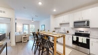 This single-floor residence features a bright, open-concept layout that seamlessly connects the kitchen, dining, and living areas. The kitchen is well-appointed with white shaker-style cabinetry extending to the ceiling, granite countertops, a white subway tile backsplash, and a full suite of stainless steel appliances, including a refrigerator, electric range, microwave, and dishwasher. A freestanding wood bar-height table with black spindle barstools serves as a natural divider between the kitchen and living space, offering a casual dining option.

The living room features light wood-look flooring, a ceiling fan, and a glass-panel door leading to a private balcony with outdoor views. The bedroom is carpeted and well-lit, equipped with a ceiling fan and a window with natural light. The bathroom mirrors the kitchen's finishes, with granite countertops, white cabinetry, and wood-look flooring. A dedicated laundry area houses a full-size Whirlpool top-load washer and dryer with wire shelving above. The overall aesthetic is clean and contemporary, with neutral whites and warm wood tones throughout.