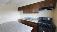This single-floor unit offers a well-organized layout designed to maximize functionality within a compact footprint. The kitchen is a standout feature, showcasing rich, dark walnut-toned cabinetry with raised-panel detailing that extends to the upper and lower storage areas, providing ample cabinet space. Light gray speckled countertops run along the workspace and extend to an adjacent breakfast bar or peninsula, adding additional prep and dining surface area. A double-basin stainless steel sink is centrally positioned beneath the upper cabinetry, while a black gas range with an overhead exhaust hood completes the cooking station. The neutral cream-toned walls throughout create a clean, understated backdrop that complements the darker cabinetry. The single-floor design ensures easy accessibility with no stairs between living areas. The bedroom and full bathroom round out the unit, offering essential living accommodations. Overall, the space presents a practical and efficiently designed interior, suitable for those seeking a streamlined, low-maintenance living environment with key kitchen amenities already in place.