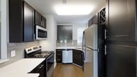 This single-floor residence offers a thoughtfully designed layout with a cohesive neutral color palette throughout. The kitchen is a standout feature, showcasing floor-to-ceiling dark espresso cabinetry that contrasts sharply with light countertops and a reflective subway tile backsplash. Stainless steel appliances, including a refrigerator, dishwasher, microwave, and electric range, provide a modern, functional workspace. A bright overhead light panel illuminates the galley-style kitchen, which also benefits from a window that invites natural light.

Adjacent to the kitchen, a white breakfast bar serves as a transition point into the living area, which features wood-style flooring, a ceiling fan, and a window overlooking outdoor greenery. The textured ceiling adds subtle architectural detail to the space.

A paneled white door leads to the bathroom, which is equipped with dark vanity cabinetry, a light countertop, and a well-lit vanity mirror. The combination of dark and light tones used throughout the residence creates a balanced, contemporary aesthetic that is both practical and visually appealing.