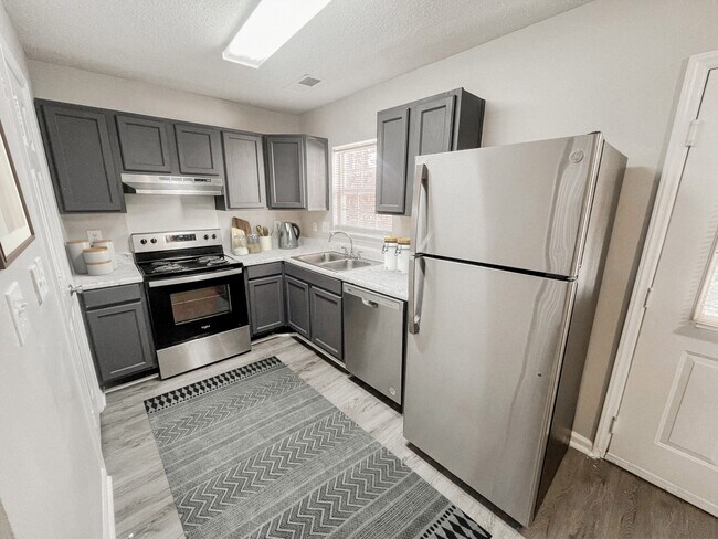 Cocina con electrodomésticos de acero inoxidable, gabinetes grises, detalles decorativos, alfombra estampada y encimeras estilo mármol. - Pendleton Townhomes