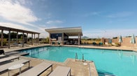 This single-story community clubhouse offers a thoughtfully designed gathering space that seamlessly blends indoor comfort with outdoor leisure. The grand entrance welcomes visitors into a bright, airy lobby featuring soaring ceilings, a statement chandelier, and large dark-framed glass doors that flood the space with natural light. The interior lounge area is appointed with neutral-toned sofas, wingback chairs, and warm accent pillows, creating a relaxed yet refined atmosphere. A striking floor-to-ceiling stone fireplace with a rustic wood mantle serves as the centerpiece of the lounge, adding warmth and architectural character. Multiple French-style glass doors connect the interior to the outdoors, reinforcing a sense of openness throughout. Outside, the resort-style pool is the highlight of the outdoor amenity area, featuring sparkling blue water, a wide surrounding deck with lounge chairs, and a modern covered cabana pavilion. The cabana is outfitted with recessed lighting, comfortable seating, and open-air access, making it ideal for relaxation and social gatherings. The fenced outdoor space offers open views of the surrounding landscape beneath expansive skies, completing the resort-inspired experience of this well-appointed community facility.