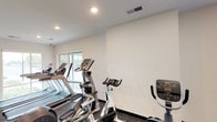 This single-level fitness space is designed to support a comprehensive cardio workout routine. The room features a clean, minimalist aesthetic with crisp white walls and a neutral-toned ceiling equipped with recessed lighting, ensuring the space is consistently and evenly illuminated. Dark baseboards provide a subtle contrast to the light walls, giving the room a polished, intentional finish.

The flooring consists of dark, textured material well-suited for high-traffic exercise use, offering both durability and grip underfoot. A variety of cardio equipment is arranged efficiently throughout the space, including multiple treadmills, elliptical trainers, Spinning-brand stationary bikes, and an upright exercise bike — providing users with a well-rounded selection of aerobic workout options.

A window along one wall allows natural light to filter into the space, contributing to an open and energizing atmosphere. Ventilation is addressed through a ceiling-mounted HVAC vent, supporting comfort during active use. The overall layout is organized and functional, with equipment positioned to allow reasonable circulation between machines. The space projects a practical, no-frills environment focused entirely on fitness utility.