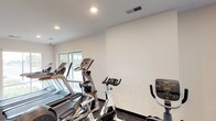 This single-level fitness space is designed to support a comprehensive cardio workout routine. The room features a clean, minimalist aesthetic with crisp white walls and a neutral-toned ceiling equipped with recessed lighting, ensuring the space is consistently and evenly illuminated. Dark baseboards provide a subtle contrast to the light walls, giving the room a polished, intentional finish.

The flooring consists of dark, textured material well-suited for high-traffic exercise use, offering both durability and grip underfoot. A variety of cardio equipment is arranged efficiently throughout the space, including multiple treadmills, elliptical trainers, Spinning-brand stationary bikes, and an upright exercise bike — providing users with a well-rounded selection of aerobic workout options.

A window along one wall allows natural light to filter into the space, contributing to an open and energizing atmosphere. Ventilation is addressed through a ceiling-mounted HVAC vent, supporting comfort during active use. The overall layout is organized and functional, with equipment positioned to allow reasonable circulation between machines. The space projects a practical, no-frills environment focused entirely on fitness utility.