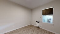 Located at 1831-1835 Harmon St in Berkeley, CA, this single-room space offers a clean, minimalist design that maximizes its compact footprint. The room features light wood-look luxury vinyl flooring throughout, lending warmth and a modern aesthetic to the interior. Neutral, light gray-beige walls create a bright and airy atmosphere, complemented by crisp white baseboards that frame the space neatly.

A wall-mounted heater provides direct, efficient heating and is positioned alongside a window fitted with a roller shade, allowing for natural light while maintaining privacy. The smooth, flat ceiling contributes to the room's clean lines and uncluttered feel.

A two-panel gray interior door with a sleek hardware finish opens to an adjacent hallway or shared area, visible in several of the images, which features tile flooring in a soft gray tone and decorative wall art — suggesting a well-maintained shared corridor. The transition between spaces is well-defined, with thoughtful material choices distinguishing the bedroom from the common area.

Overall, this space presents a straightforward, functional design that is well-suited for single occupancy. Its fresh finishes and practical amenities reflect a carefully maintained property in a prime Berkeley location.