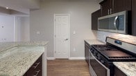This two-floor space offers a well-designed layout with thoughtful finishes throughout. The main living area is characterized by warm hardwood-style flooring, decorative white columns, and a combination of track lighting and a ceiling fan, creating a bright and functional atmosphere. A window allows natural light to fill the space, enhancing its open feel. The upper level connects to the lower floor via a staircase visible in the architectural overview.

The kitchen is a standout feature, equipped with rich dark cabinetry, granite countertops with subtle veining, a stone tile backsplash, and a full suite of stainless steel appliances including a gas range, microwave, refrigerator, and dishwasher. A double-basin sink with modern fixtures completes the well-appointed cooking space.

The bathrooms continue the refined aesthetic, featuring granite vanity tops, dark wood cabinetry with brushed nickel hardware, and large mirrors with multi-bulb lighting. One bathroom includes a tub-shower combination with wood-look flooring.

Bedrooms are carpeted and receive natural light through blinds-fitted windows. The attached garage provides additional utility, featuring a textured concrete floor and an automated door opener. Throughout, neutral wall tones and consistent finishes create a cohesive, modern ambiance.