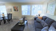This single-floor space offers a comfortable and well-appointed living environment with a warm, inviting ambiance. The living room serves as the centerpiece of the space, featuring plush wall-to-wall carpeting that adds softness underfoot and contributes to the room's cozy atmosphere. A pair of large windows floods the room with abundant natural light while framing views of lush, mature trees outside, creating a tranquil connection to the outdoors. Baseboard heating runs along the perimeter of the room, ensuring year-round comfort. The space is tastefully styled with a dark-framed sofa accented by patterned and solid throw pillows, complemented by a sleek black coffee table topped with decorative accents. A framed abstract artwork adds a pop of color to the neutral, light-toned walls, enhancing the room's aesthetic appeal. A classic table lamp provides warm supplemental lighting in the corner. The overall design balances practicality with style, making this a functional yet charming living space ideal for relaxing or entertaining.