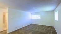 This single-story space presents a straightforward, functional layout centered around a generously sized room visible in the image. The room features neutral white walls that contribute to a bright and open atmosphere, complemented by carpeted flooring in a muted, earthy tone. A textured popcorn ceiling adds a classic touch overhead, while floor-level vents indicate a built-in climate control system for year-round comfort. A single window along the far wall invites natural light into the space, offering a view of mature outdoor greenery. The clean, minimalist design of the room provides a versatile canvas suitable for a variety of uses. White baseboards frame the perimeter of the room, and standard electrical outlets are conveniently positioned along the walls. The overall ambiance is understated and practical, with the neutral color palette and carpeted floor lending a quiet, comfortable quality to the interior. The absence of clutter and the simplicity of the finishes make this a highly adaptable space.
