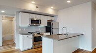 This thoughtfully designed single-floor residence combines modern aesthetics with functional living spaces. The open-concept kitchen is a standout feature, showcasing white shaker cabinetry, a herringbone-patterned tile backsplash, stainless steel appliances including a range, microwave, and refrigerator, and a central island with a grey quartz countertop and undermount sink — ideal for both cooking and casual dining. Rich, warm-toned hardwood flooring flows throughout the kitchen and living areas, complemented by recessed lighting that creates a bright and welcoming ambiance.

The living area is generously proportioned, with large floor-to-ceiling windows framing expansive urban skyline views, flooding the space with natural light. The bedrooms continue the theme of abundant natural light, featuring oversized windows with city views, soft carpeting, and ample closet space with sliding doors.

The two bathrooms are finished with a clean, contemporary palette. One features a walk-in glass-enclosed shower with marble-look tile surround and matte black fixtures, while the second offers a full soaking tub with a matching marble-look tile surround. Both bathrooms include white vanities, white countertops, and warm-toned tile flooring, delivering a polished and cohesive aesthetic throughout.