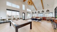 Located in Thornton, Colorado, this contemporary community clubroom is a well-appointed amenity space designed for socializing and recreation. The room is defined by its dramatic vaulted ceiling with exposed natural wood beams that draw the eye upward, creating an expansive, airy atmosphere throughout the open-plan layout.

The space is anchored by a sleek, dark-framed pool table positioned beneath a striking gold starburst chandelier — a bold focal point that adds a touch of elegance to the recreational area. Light hardwood flooring runs continuously throughout, lending warmth and visual cohesion to the entire space.

A generous seating and dining area occupies one side of the room, furnished with a mix of communal tables, navy upholstered chairs, and continuous leather banquette seating along the windowed wall. Clusters of modern black wire pendant lights hang above, providing layered illumination while reinforcing the contemporary aesthetic.

A bar-height counter with leather-and-metal stools sits adjacent to a dramatic steel-framed glass partition wall, offering a more casual perch and a view into an adjoining space. Along another wall, a standing-height counter with additional bar seating provides direct access to the exterior via a glass door.

Large black-trimmed windows line multiple walls, flooding the interior with natural light and offering views of the outdoor pool area and surrounding landscape — seamlessly connecting the indoors with the outdoors.