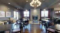 This single-floor common lounge is a well-appointed gathering space designed to balance comfort and functionality. The room makes an immediate impression with its ornate wrought-iron chandelier fitted with drum shades, suspended from a vaulted ceiling that lends a sense of vertical grandeur. Rich, dark hardwood floors run throughout, grounding the space and complementing the overall warm yet sophisticated aesthetic.

A textured tile fireplace serves as the room's focal point, flanked by burgundy drape-accented windows that frame natural light from multiple sides of the space. A neutral-toned area rug and a glass coffee table anchor a comfortable seating arrangement of cream upholstered sofas and accent chairs positioned around the hearth.

Supplementing the lounge seating, the space also incorporates several dark-wood bistro-style tables and chairs, offering a more structured setting suitable for casual dining or work. Recessed lighting throughout ensures even illumination alongside the natural light provided by the generous window placement. Glass-paneled doors on one side offer access to an adjacent outdoor area, further enhancing the sense of openness within this versatile, elegantly designed space.