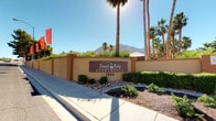 Desert Ridge Apartments is a well-appointed residential community that makes a strong first impression with its distinctive stucco perimeter wall, manicured desert landscaping, and tall swaying palm trees framing the entrance signage. The community is set against a dramatic backdrop of rugged mountain ranges visible beneath wide, sun-drenched skies, lending the property a striking sense of place within its desert environment.

At the heart of the community lies a dedicated playground area, thoughtfully designed for outdoor recreation. The play structure features climbing frames, slides, and decorative leaf-canopy toppers, all set within a generous sand surface for safe play. Mature shade trees surround the area, offering relief from the desert sun and creating a comfortable, inviting atmosphere for residents.

The surrounding grounds are lush with green grass and mature trees, contrasting beautifully with the warm earth-toned stucco exteriors of the multi-story residential buildings nearby. Concrete walkways wind through the community, connecting the various amenity areas. Clear posted signage reinforces community standards, reflecting an attentive and organized management approach. Overall, Desert Ridge Apartments combines practical amenities with natural desert beauty to create a vibrant and livable community environment.