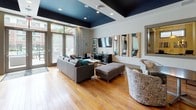 This single-floor communal space offers a sophisticated and well-appointed interior designed for both relaxation and social interaction. The most striking architectural feature is the bold navy blue ceiling, which contrasts dramatically with the bright white walls and creates a sense of defined zones throughout the open layout. Warm hardwood flooring flows continuously across the entire space, lending a cohesive and inviting atmosphere.

The main lounge area is furnished with a generous sectional sofa, ottoman seating, and accent chairs arranged around a central coffee table, with a large wall-mounted flat-screen television serving as a focal point. Floor-to-ceiling windows along one wall allow abundant natural light to pour in, offering views of an outdoor area beyond.

A secondary seating zone features additional lounge chairs and mirrored wall accents, enhancing the sense of openness. Toward the rear, a dedicated dining and wet bar area includes a dark-topped communal table with leather chairs, a sleek bar counter with bar stools, a sink, and tiled backsplash, making it well-suited for gatherings. Glass-paneled interior doors connect to adjacent rooms, adding transparency and visual depth throughout.