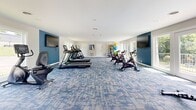 Located at 2411 Pulaski Hwy in Columbia, Tennessee, this fitness center offers a spacious, single-floor layout designed to support a comprehensive workout experience. The interior is finished with a distinctive blue and gray patterned carpet that adds a modern aesthetic throughout the open exercise area. Walls are painted in a clean combination of crisp white and bold accent blue, contributing to an energetic yet welcoming atmosphere.

The facility is outfitted with a diverse array of cardio equipment, including multiple treadmills, elliptical cross-trainers, and stationary cycling bikes positioned to make the most of the available floor space. A dedicated free weight and dumbbell rack rounds out the strength training options. Multiple wall-mounted flat-screen televisions are strategically placed throughout the room, ensuring visibility from virtually any workout station.

Natural light flows generously into the space through large windows and French-style glass doors positioned along multiple walls. These doors open directly onto an outdoor balcony or terrace area, providing views of the surrounding greenery and a seamless connection to the outdoors. Recessed and flush-mount ceiling lighting supplements natural daylight, keeping the space bright at all hours. The overall design prioritizes both functionality and comfort, making it a practical and well-appointed fitness amenity.