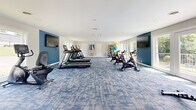 Located at 2411 Pulaski Hwy in Columbia, Tennessee, this fitness center offers a spacious, single-floor layout designed to support a comprehensive workout experience. The interior is finished with a distinctive blue and gray patterned carpet that adds a modern aesthetic throughout the open exercise area. Walls are painted in a clean combination of crisp white and bold accent blue, contributing to an energetic yet welcoming atmosphere.

The facility is outfitted with a diverse array of cardio equipment, including multiple treadmills, elliptical cross-trainers, and stationary cycling bikes positioned to make the most of the available floor space. A dedicated free weight and dumbbell rack rounds out the strength training options. Multiple wall-mounted flat-screen televisions are strategically placed throughout the room, ensuring visibility from virtually any workout station.

Natural light flows generously into the space through large windows and French-style glass doors positioned along multiple walls. These doors open directly onto an outdoor balcony or terrace area, providing views of the surrounding greenery and a seamless connection to the outdoors. Recessed and flush-mount ceiling lighting supplements natural daylight, keeping the space bright at all hours. The overall design prioritizes both functionality and comfort, making it a practical and well-appointed fitness amenity.