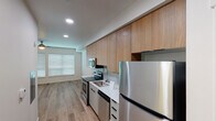This contemporary single-floor unit offers a clean, modern aesthetic throughout, highlighted by warm light oak hardwood flooring and crisp white walls that create a bright, welcoming atmosphere. The open-concept kitchen is well-appointed with warm wood upper cabinetry extending to the ceiling, light gray lower cabinets, white quartz countertops, a subway tile backsplash, and a full suite of stainless steel appliances including a refrigerator, electric range, built-in microwave, and dishwasher. Recessed lighting illuminates the kitchen efficiently. The adjoining living area is generously proportioned, bathed in natural light from large windows dressed with horizontal blinds, and features a ceiling fan for added comfort. The bedroom provides ample storage with two side-by-side closets outfitted with built-in shelving and rod systems, offering well-organized space for clothing and personal items. The bathroom presents a clean, minimalist design with a wide white quartz vanity, an undermount sink, a large mirror, and contemporary light fixture. Consistent hardwood flooring flows throughout the unit, tying together the cohesive modern design. The overall layout is functional and thoughtfully designed, making efficient use of the available space.