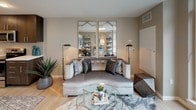 This single-floor, one-bedroom, one-bathroom residence features an open-concept layout that maximizes the sense of space throughout. The living area is finished with light hardwood-style flooring and neutral warm-gray walls, creating a versatile backdrop. A curved sofa anchors the living space beneath decorative framed mirrors, while floor lamps add ambient lighting.

The kitchen is outfitted with dark espresso cabinetry, both upper and lower, complemented by open shelving and a textured tile backsplash. Stainless steel appliances — including a range, built-in microwave, and dishwasher — line the countertop, which features a clean white surface and an undermount sink.

The open bedroom area continues the neutral color palette with gray walls and light flooring, offering a cozy, private retreat with ample room for furnishings.

The bathroom showcases tile flooring, a white vanity with light wood cabinetry, and a shower-tub combination with a dark curtain. Framed abstract artwork and teal accent towels add a pop of color against the gray walls, tying the overall design aesthetic together cohesively throughout the residence.