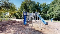 This outdoor playground offers an engaging and well-equipped recreational space designed for active play. The centerpiece is a multi-level play structure finished in blue and silver, featuring two prominent slides — one vertical blue slide and one arching blue slide with a curved metal frame — along with climbing ladders and raised platforms with safety railings. The structure is built to encourage a range of physical activities, from climbing and sliding to imaginative play across its connected levels.

The ground surface throughout the play area is covered in a generous layer of wood chip mulch, providing a natural, impact-absorbing safety surface beneath all the equipment. The perimeter of the space is defined by mature deciduous trees, their full green canopies offering natural shade and a pleasant, park-like atmosphere. The open sky above allows for abundant natural light, creating a bright and inviting environment on clear days.

The overall setting feels spacious and well-maintained, with a clean layout that allows easy visibility across the entire play area. The combination of durable metal equipment, natural surfacing, and a lush tree backdrop gives the space a welcoming, community-oriented character suited for children of various ages.