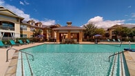 Located at 7145 Reading Rd in Rosenberg, Texas, this outdoor amenity space centers around a generously sized resort-style swimming pool with crystal-clear blue water. The pool area is a standout feature of the community, designed to offer both relaxation and recreation under the expansive Texas sky. At the far end of the pool, a classically styled covered cabana pavilion with a distinctive hip roof and cupola serves as a focal point, complete with a decorative water feature that adds both visual appeal and ambient sound to the environment. The surrounding pool deck is finished in a warm, neutral tone and is well-equipped with lounging chairs and umbrella-shaded seating areas, providing ample space for sunbathing and outdoor gatherings. Lush, manicured landscaping lines the perimeter, adding a touch of greenery that complements the warm earth-toned architecture of the multi-story residential buildings visible in the background. A gated entry ensures privacy and safety, with clearly posted pool policies at the entrance. The overall ambiance is that of a well-maintained, community-oriented outdoor retreat that balances practical amenities with an inviting, resort-inspired aesthetic.