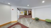 This single-floor communal space functions as a welcoming lobby and lounge area defined by a thoughtful blend of materials and color. The entry foyer features a tall, light-filled interior with a set of wood-trimmed steps leading to a glass-paneled door, flanked by a bank of built-in storage lockers. Polished concrete flooring extends throughout much of the space, contrasted by patterned area rugs in deep red and neutral tones that delineate seating zones.

The central lounge area is anchored by a floor-to-ceiling tile-clad fireplace, which serves as a striking architectural focal point. Surrounding it is an arrangement of upholstered armchairs in patterned and solid fabrics, complemented by rounded ottomans in olive green. A large, vibrant floral mural in warm yellows and blues adds a bold artistic element to one wall, contributing to the space's energetic character.

Accent walls painted in yellow-green provide warmth and visual continuity throughout. Large windows along one side allow natural light to fill the space while offering views of outdoor landscaping. Additional corridors branch off toward mailboxes and interior hallways, suggesting this serves as a central hub for a larger residential or institutional building. Recessed lighting throughout ensures the space remains well-lit and inviting.