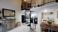 This two-story space offers a thoughtfully designed layout that maximizes both functionality and visual interest. The centerpiece of the main level is a bold black spiral staircase, which serves as a striking architectural focal point while efficiently connecting the two floors. The open-concept main level features a carpeted living area with warm, neutral-toned walls and ample room for comfortable furnishings. A decorative console table and mirror accent the entryway, adding a welcoming touch. The kitchen is well-appointed with white cabinetry, black appliances including a refrigerator and electric range, and dark countertops, creating a classic contrast. A dining area with traditional wooden chairs sits adjacent to the kitchen, making the space ideal for everyday living and entertaining. The upper loft level, visible from below through black metal railings, provides an open and airy feel while offering additional functional space. High ceilings on the main level enhance the sense of openness and allow natural sightlines to the loft above. Light wood-style flooring in the kitchen transitions smoothly to carpet in the living areas, adding warmth and texture throughout this well-connected, vertically expansive home.
