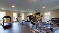 This dedicated fitness space is thoughtfully designed to support a comprehensive workout routine. The room is finished with clean white walls and a smooth grey ceiling outfitted with recessed lighting, ensuring even illumination throughout the space. Multiple large windows are positioned along the walls, allowing ample natural light to fill the room while offering views of the surrounding greenery outdoors.

The flooring consists of a durable, dark rubberized surface well-suited for exercise equipment and high-traffic use. The room is furnished with an impressive array of cardio equipment, including a rowing machine, a treadmill, and multiple elliptical trainers, providing a variety of workout options. A wall-mounted flat-screen television is centrally positioned, allowing users to stay entertained during their sessions.

The layout of the equipment is organized to maximize usable floor space while maintaining clear pathways between machines. The overall ambiance is clean, modern, and purpose-driven, making it a highly functional environment for fitness enthusiasts seeking a private, well-appointed space to exercise.