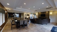 This well-appointed single-floor space showcases an open-concept layout that seamlessly integrates a fully equipped kitchen, a generous dining area, and a relaxed lounge zone. The kitchen is outfitted with dark cabinetry, stainless steel appliances including double wall ovens, and a prominent overhead range hood, making it well-suited for preparing meals for a crowd. A large center island provides additional workspace and casual seating.

The dining area accommodates an expansive dark-wood table surrounded by upholstered high-back chairs, offering ample seating for group gatherings. Natural stone tile flooring in a neutral palette flows throughout the space, lending a clean and cohesive aesthetic.

The lounge area is anchored by a large flat-screen television mounted on a striking stacked-stone accent wall, adding a warm, textural focal point. Comfortable sectional seating and tasteful decorative lighting complete this relaxed setting. Recessed lighting is installed throughout, complemented by a tray ceiling detail that adds architectural depth overhead. A console table with decorative lamps and artwork provides elegant transitional elements between the dining and lounge areas. Overall, the space presents a refined yet inviting atmosphere ideal for communal use and entertaining.