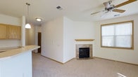 This single-floor space offers a clean, functional layout characterized by neutral white walls, warm wood trim, and plush carpeting throughout. The main living area is anchored by a tile-surround fireplace with a wood mantel, serving as a natural focal point. A ceiling fan overhead provides practical climate control, while a window fitted with blinds allows natural light to filter into the room. The open layout connects the living area to an adjacent space, partially visible behind a dividing wall, suggesting a seamless flow between the living and dining or kitchen zones. A countertop ledge is visible to one side, hinting at a breakfast bar or pass-through kitchen feature. The space maintains a bright, airy feel through its consistent white palette and straightforward design. Recessed ceiling vents and flush-mount lighting contribute to the practical, well-maintained character of the interior. Hallway access visible in the background indicates additional rooms beyond the main living space. Overall, the layout is efficient and inviting, with the fireplace and natural light adding warmth and comfort.