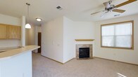 This single-floor space offers a clean, functional layout characterized by neutral white walls, warm wood trim, and plush carpeting throughout. The main living area is anchored by a tile-surround fireplace with a wood mantel, serving as a natural focal point. A ceiling fan overhead provides practical climate control, while a window fitted with blinds allows natural light to filter into the room. The open layout connects the living area to an adjacent space, partially visible behind a dividing wall, suggesting a seamless flow between the living and dining or kitchen zones. A countertop ledge is visible to one side, hinting at a breakfast bar or pass-through kitchen feature. The space maintains a bright, airy feel through its consistent white palette and straightforward design. Recessed ceiling vents and flush-mount lighting contribute to the practical, well-maintained character of the interior. Hallway access visible in the background indicates additional rooms beyond the main living space. Overall, the layout is efficient and inviting, with the fireplace and natural light adding warmth and comfort.