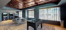 This well-appointed single-floor amenity space is designed for entertainment and socializing, combining thoughtful design with a rich variety of recreational features. The interior is anchored by a distinctive coffered wood ceiling that adds warmth and a sense of architectural sophistication throughout the main area. Light hardwood flooring runs underfoot, complemented by layered area rugs that define distinct activity zones.

The game room serves as the centerpiece, housing a full-size pool table with a classic red felt surface, a long shuffleboard table, a foosball table, and a vintage arcade cabinet — offering a comprehensive range of entertainment options. A dedicated lounge area features comfortable seating arranged around a wall-mounted television, creating a relaxed gathering spot. Large windows dressed with wood-slat blinds flood the space with natural light and provide views of a waterway and outdoor seating area beyond.

An adjacent wet bar area is fitted with warm wood cabinetry, open shelving set against exposed brick, and under-counter refrigeration — ideal for hosting. A keypad-secured entry door provides controlled access to the space. Black-framed glass partition doors connect the main game area to additional rooms, adding an open yet defined layout. The overall ambiance is modern and moody, with deep teal walls, dark architectural accents, and warm wood tones creating an inviting, upscale atmosphere.