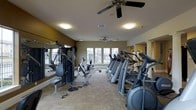 This fitness center offers a comprehensive workout environment designed to accommodate a range of exercise routines. The space is outfitted with a row of cardio machines, including treadmills and ellipticals, positioned along one side of the room for efficient use of floor space. A multi-station strength training machine and free weights are available on the opposing side, catering to resistance and weight training needs. Large windows along the far wall invite natural light into the space, creating a bright and energizing atmosphere. Full-length mirrors are mounted along one wall, enhancing the sense of openness while allowing users to monitor their form during workouts. The light-toned flooring adds to the airy feel of the room. Two ceiling fans are installed overhead, providing consistent air circulation throughout the space to maintain comfort during intense exercise sessions. Decorative columns near the rear of the room add a subtle architectural detail. The warm-toned walls complement the overall design, resulting in a functional yet inviting fitness facility suitable for a variety of workout preferences.