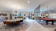 This single-floor amenity lounge is thoughtfully designed to offer a dynamic mix of social, recreational, and relaxation spaces. The main gathering area is anchored by a large round dining table set atop a bold circular area rug, illuminated by a striking sputnik-style chandelier that serves as a focal point of the room. Light wood-look flooring flows throughout, lending warmth and continuity across the space.

A sheer curtain partition elegantly divides the main lounge from a dedicated game area, which features a full-size pool table with a vibrant red felt surface, set beneath its own pendant lighting. Direct access to an outdoor patio through glass double doors floods the interior with natural light and provides a seamless indoor-outdoor connection.

The kitchenette is outfitted with wood-grain cabinetry, crisp white quartz countertops, stainless steel appliances including a microwave and beverage refrigerator, and a functional sink — ideal for catering and entertaining. An artistically carved white decorative panel adds a sculptural element to this area.

Lounge seating nooks are furnished with warm brown accent chairs, industrial-style side tables, and decorative wall sconces. Textured wallcoverings, curated artwork, and thoughtful lighting throughout give the space a refined yet welcoming ambiance suited for both casual gatherings and private events.