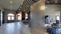 This single-floor space makes an immediate impression with its striking interior design and thoughtful architectural details. The entrance area is defined by dramatic semi-transparent partition panels adorned with intricate gold branch patterns, creating a visually captivating threshold that separates zones while maintaining an open, airy feel throughout.

The ceiling is a standout feature, showcasing bold black-and-white diagonal stripes that draw the eye upward and add a sense of dynamic energy to the space. Recessed track lighting is embedded throughout, providing focused illumination that complements the overall ambiance. A decorative chandelier adds an elegant touch to the lounge area beyond.

The main lounge area is anchored by comfortable gray upholstered seating arranged around a central gathering space, set atop light wood-tone flooring that runs seamlessly throughout. A warm, earthy accent wall in deep terracotta contrasts beautifully with the cooler tones elsewhere, while tall windows dressed with rust-colored drapes allow ample natural light to filter in and frame views of the exterior. A fireplace further enhances the welcoming atmosphere of the lounge. The overall design balances bold decorative elements with functional, open living spaces, resulting in a sophisticated and memorable interior environment.