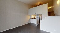 This two-story space makes a striking first impression with its dramatic double-height ceilings in the main living area, where the open vertical expanse creates a sense of grandeur. The ground floor is finished with dark, wood-look tile flooring throughout, lending a sleek and modern aesthetic. Large vertical blinds cover a floor-to-ceiling window, flooding the entry area with natural light while offering privacy and light control.

The kitchen is thoughtfully designed with crisp white shaker-style cabinetry, dark countertops, and a tile backsplash. Stainless steel appliances — including an electric range, over-the-range microwave, and refrigerator — complete the functional and stylish cooking space. A pass-through counter connects the kitchen to the main living area, maintaining an open feel.

A staircase leads to the upper level, where the design takes on a cozier character with wall-to-wall carpeting, textured walls, and ceiling fans in each room. The upper floor features sloped ceilings that follow the roofline, adding architectural interest and a distinctive loft-like quality. Open railings along the upper hallway overlook the living area below, reinforcing the home's open and connected layout. Each upper-level room benefits from natural light through vertical-blind-covered windows.