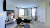 This single-floor space offers a comfortable and thoughtfully designed living environment centered around a spacious living room. The room is bathed in natural light streaming through multiple large windows dressed with elegant patterned draping curtains, creating a warm and inviting atmosphere. Neutral wall tones and light carpeting throughout contribute to an open, airy feel. The living room is generously sized, accommodating a classic upholstered sofa with coordinating armchairs, a dark leather ottoman coffee table, and a round accent table in the foreground. Recessed ceiling lighting supplements the natural light, ensuring the space remains bright at any time of day. A tall bookcase anchors one corner of the room, adding both function and character. Decorative artwork, including a framed botanical print, adds a refined touch to the walls. The overall ambiance is clean, classic, and welcoming, reflecting a timeless interior style that balances practicality with aesthetic appeal. The layout flows naturally, making the living area feel cohesive and well-proportioned for everyday living and relaxed entertaining.