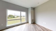 This contemporary single-floor unit is designed with a clean, modern aesthetic throughout. Light-toned hardwood flooring runs continuously across the living areas and bedroom spaces, lending warmth and cohesion to the interior. The walls are finished in a soft grey palette, creating a neutral and inviting atmosphere.

The kitchen is well-appointed with stainless steel appliances, including an electric range, over-the-range microwave, and dishwasher. Sleek grey cabinetry with brushed metal hardware pairs with a quartz-style countertop and subway tile backsplash, delivering a polished, functional workspace.

The bedroom areas are generously sized and benefit from large picture windows that frame views of surrounding green spaces and open landscape, filling the rooms with abundant natural light. Closet space is incorporated directly into the bedroom, offering practical built-in storage with a clothing rail.

The bathroom is finished with large-format tile flooring and features a grey vanity with an undermount sink and matching countertop, a toilet, and a tiled shower stall. A mirror and in-cabinet storage add to the room's functionality. An in-unit stacked washer and dryer is also present, adding everyday convenience. Overall, the space presents a bright, move-in-ready layout with contemporary finishes throughout.