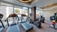 This fitness center combines thoughtful design with comprehensive workout amenities, delivering a polished and functional space for exercise. The interior is distinguished by a striking geometric-patterned accent wall in neutral tones, complemented by warm wood paneling and soft, textured carpeting throughout. Circular pendant light fixtures and recessed lighting provide a refined, contemporary atmosphere.

The cardio zone is positioned along a wall of expansive floor-to-ceiling windows, allowing abundant natural light to fill the space while offering open city views — an inspiring backdrop for treadmills and elliptical machines. Strength training is well-represented, with a selection of cable and weight machines, free weights, kettlebells, and stability balls neatly arranged throughout the facility.

A built-in dark wood shelving unit with a marble countertop and large wall-mounted mirror serves as a focal point in one section of the room, alongside a dedicated TV display. A water bottle filling station and additional mounted televisions ensure convenience during workouts. A louvered door provides access to additional facilities. The space is bright, airy, and meticulously finished, offering a premium fitness experience with an upscale, hotel-quality aesthetic.