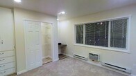 This single-floor space offers a functional and straightforward layout designed for comfortable everyday living. The main room features soft wall-to-wall carpeting underfoot, creating a warm and quiet environment. A large window fitted with horizontal blinds allows for adjustable natural light while providing a view of the surrounding outdoor greenery. Climate control is well-covered, with both a baseboard heater and a wall-mounted air conditioning unit ensuring year-round comfort. Recessed lighting in the ceiling provides clean, even illumination throughout the room. A standout feature is the walk-in closet, accessed through double bi-fold doors, which offers built-in wire shelving for organized storage across multiple levels. Adjacent to the closet entrance is a built-in desk nook set on tile flooring, creating a defined workspace or utility area separate from the carpeted main living space. The walls are finished in a neutral cream tone, contributing to a bright and airy atmosphere. The overall design is practical and efficient, making smart use of the available space with dedicated zones for rest, work, and storage all within a compact, single-level footprint.